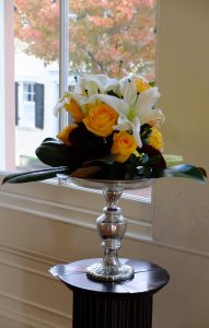 This elegant statement piece is the perfect compliment to any bride. Warm roses, pristine white lilies, magnolia and deep purple mums combine atop a mahogany pedestal.