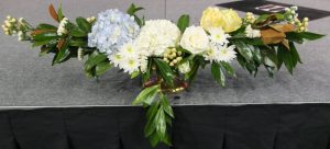 A large brass oblong footed vessel is the perfect choice for this massive stage arrangement. Shocking blue hydrangea transitions into white hydrangea then white Playa Blanca roses. Cream hypericum provides the structure to extend the arrangement well past the dimensions of the vessel. It is filled out with magnolia, white statice, and yellow Toscana roses to achieve a classic formal look.